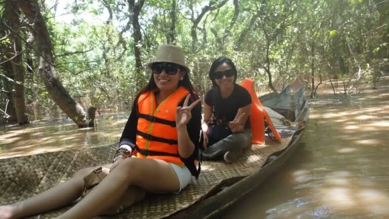 wo smiling female tourists wearing sunglasses and life jackets on a traditional wooden boat cruising through a sunlit flooded forest in Cambodia.