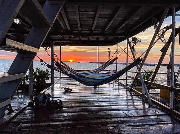 View from the wooden deck of a riverboat showing hammocks strung up against a vibrant orange sunset over a vast lake.