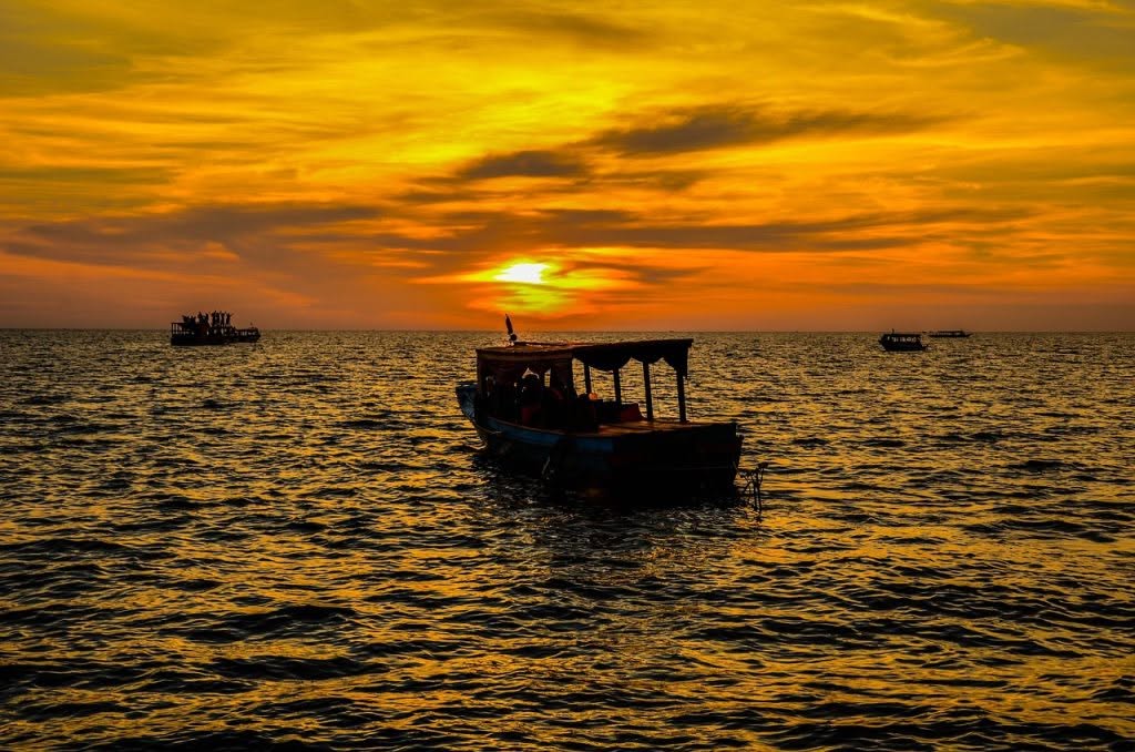 Traditional wooden tour boat carrying passengers during a vibrant golden sunset on Tonle Sap Lake in Siem Reap, Cambodia.