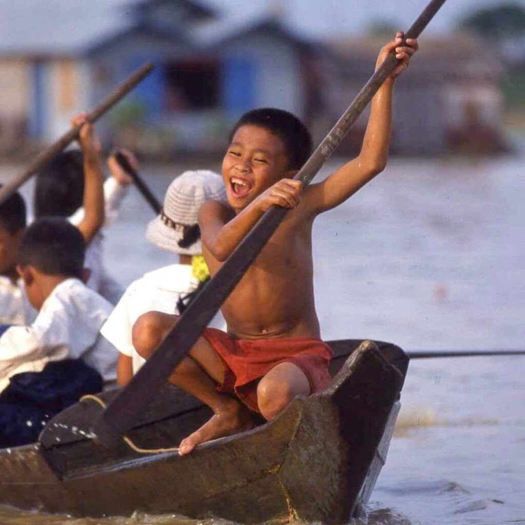 A young Cambodian boy laughing while rowing a small wooden boat through a floating village on Tonle Sap Lake.