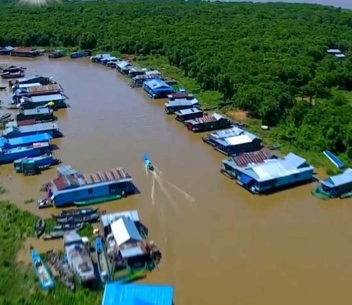 An aerial drone view of a floating village with colorful houses lining a brown river waterway surrounded by a dense green forest on a Khmer Detours tour in Cambodia.