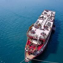 An aerial drone view of a large multi-deck riverboat mothership with a spacious top deck dining area floating on clear blue water in Cambodia during a Khmer Detours tour.