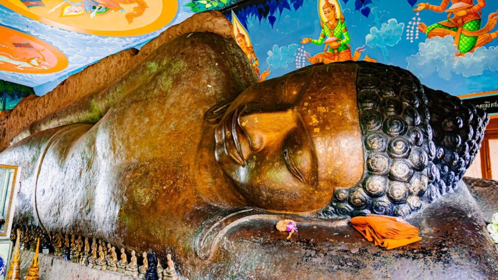A massive ancient stone reclining Buddha statue carved into a boulder at Wat Preah Ang Thom, featuring gold leaf details and a colorful painted ceiling above, as seen in image_63b690.jpg by Khmer Detours.