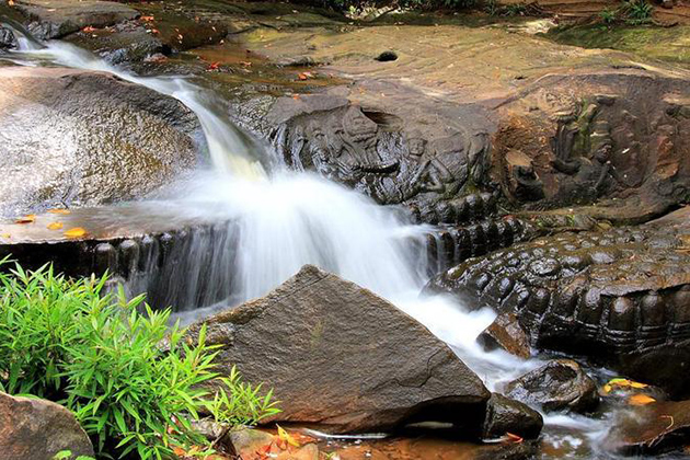 A rushing jungle waterfall flowing over ancient Hindu stone carvings on the rocky riverbed at Kbal Spean in Phnom Kulen, Cambodia, as seen in image_63581d.jpg by Khmer Detours.