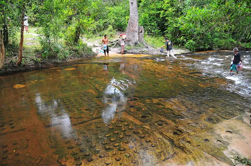 Clear water flowing over hundreds of ancient Hindu linga carvings on a rocky riverbed, with local villagers walking along the riverbank, as seen in image_635f26.jpg by Khmer Detours.
