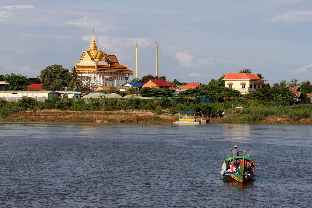 A colorful traditional Cambodian longtail boat navigating a wide river with a golden-roofed Buddhist temple (Wat) visible on the lush green riverbank, as seen in image_6348a0.jpg by Khmer Detours.