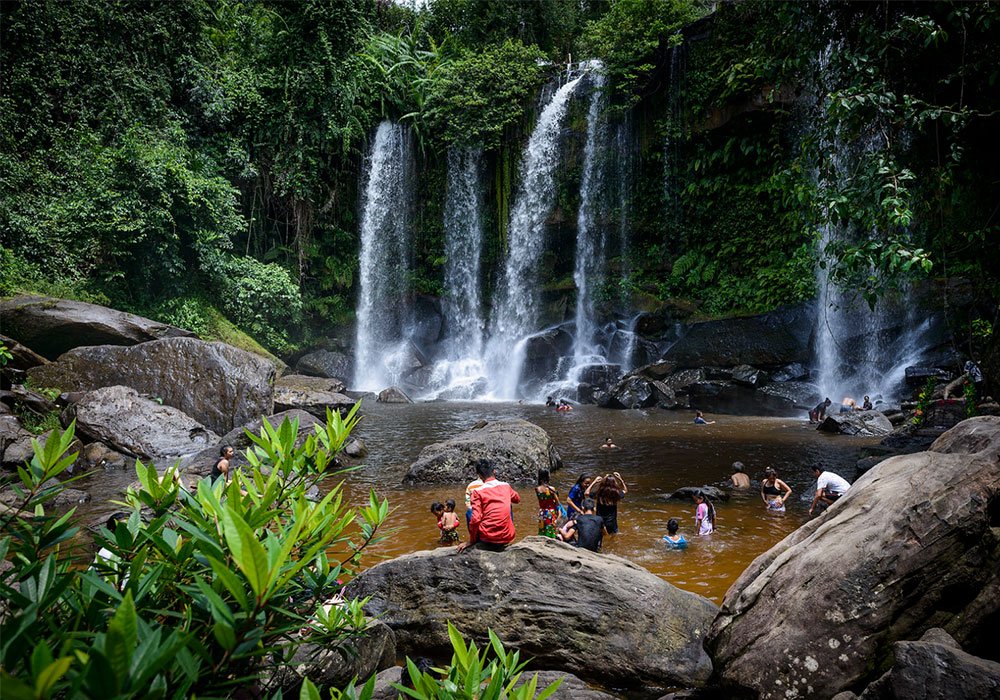 Visitors swimming and relaxing in a natural pool at the base of a beautiful multi-tiered jungle waterfall surrounded by lush green trees, as seen in image_63b1f5.jpg by Khmer Detours.