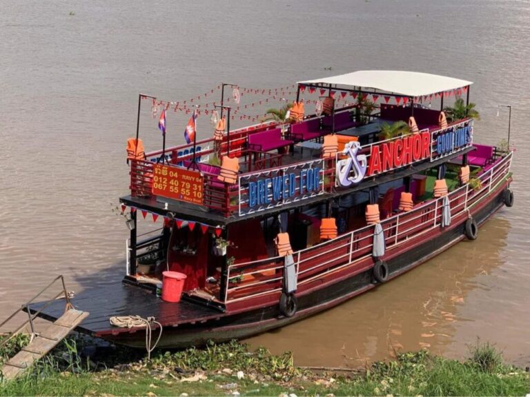 A double-decker Mekong River cruise boat named Ravy 04 docked at the riverbank, featuring Anchor Beer signage and Cambodian flags, as seen in image_6341f5.jpg by Khmer Detours.