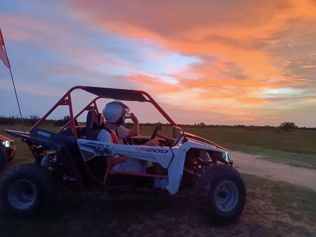 A tourist wearing a white helmet sits in an off-road buggy vehicle on a rural dirt path, taking a photo of a brilliant orange and pink sunset sky.