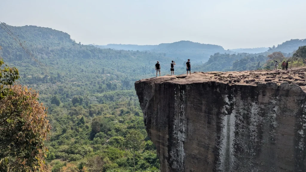 Hikers standing on the edge of a massive, sheer rock cliff overlooking a vast, dense green jungle valley under a hazy sky, as seen in image_635402.jpg by Khmer Detours.